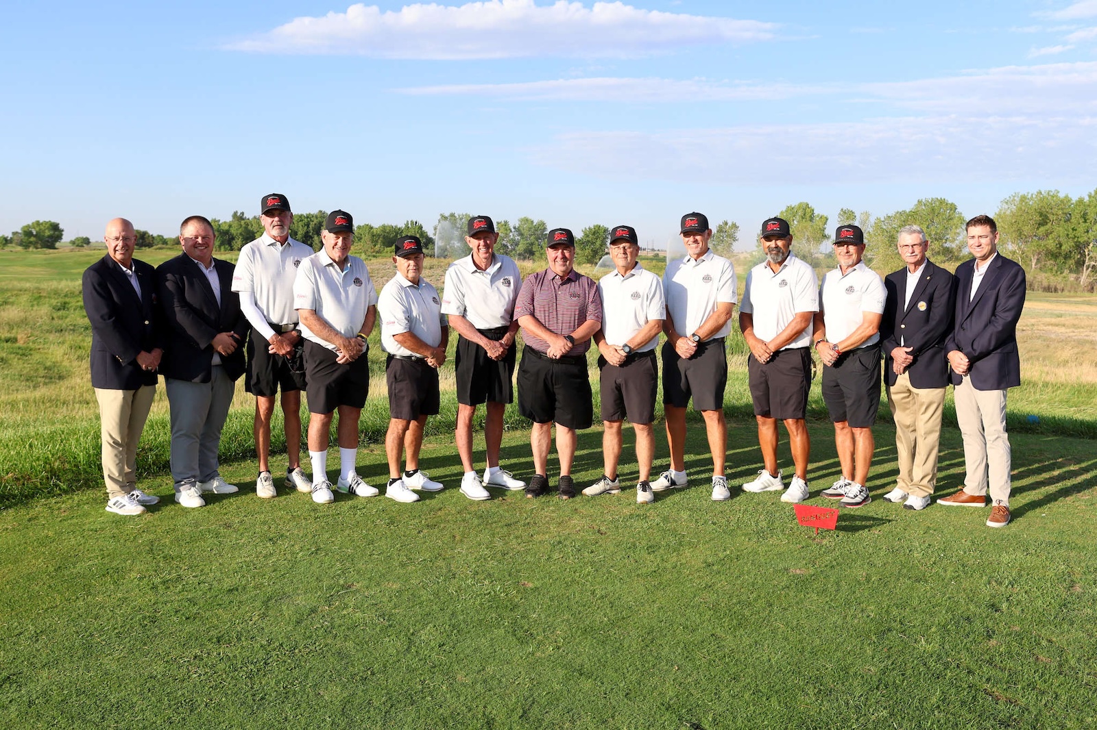 Group of about 15 men in polo shirts and caps posing for a photo on a sunny golf course nearby trees and green grass.