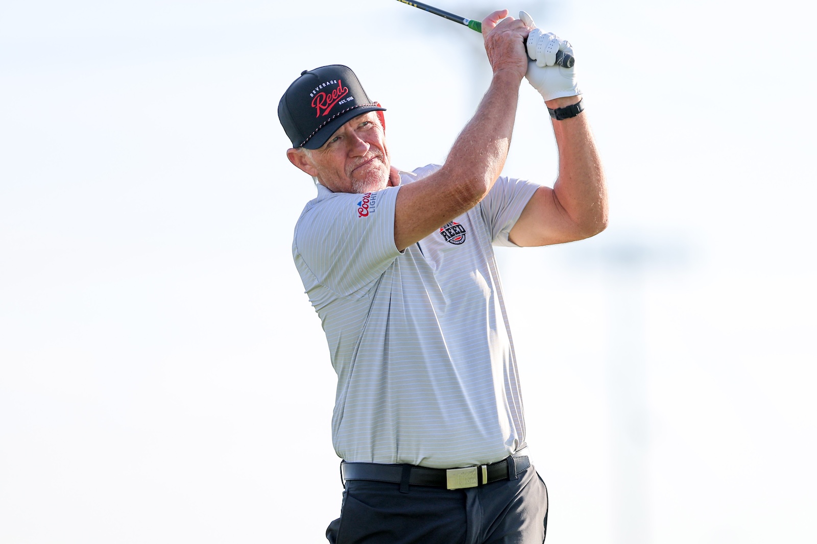 Adult male golfer mid-swing, wearing a light polo and dark cap, focusing on his follow-through against a bright sky.