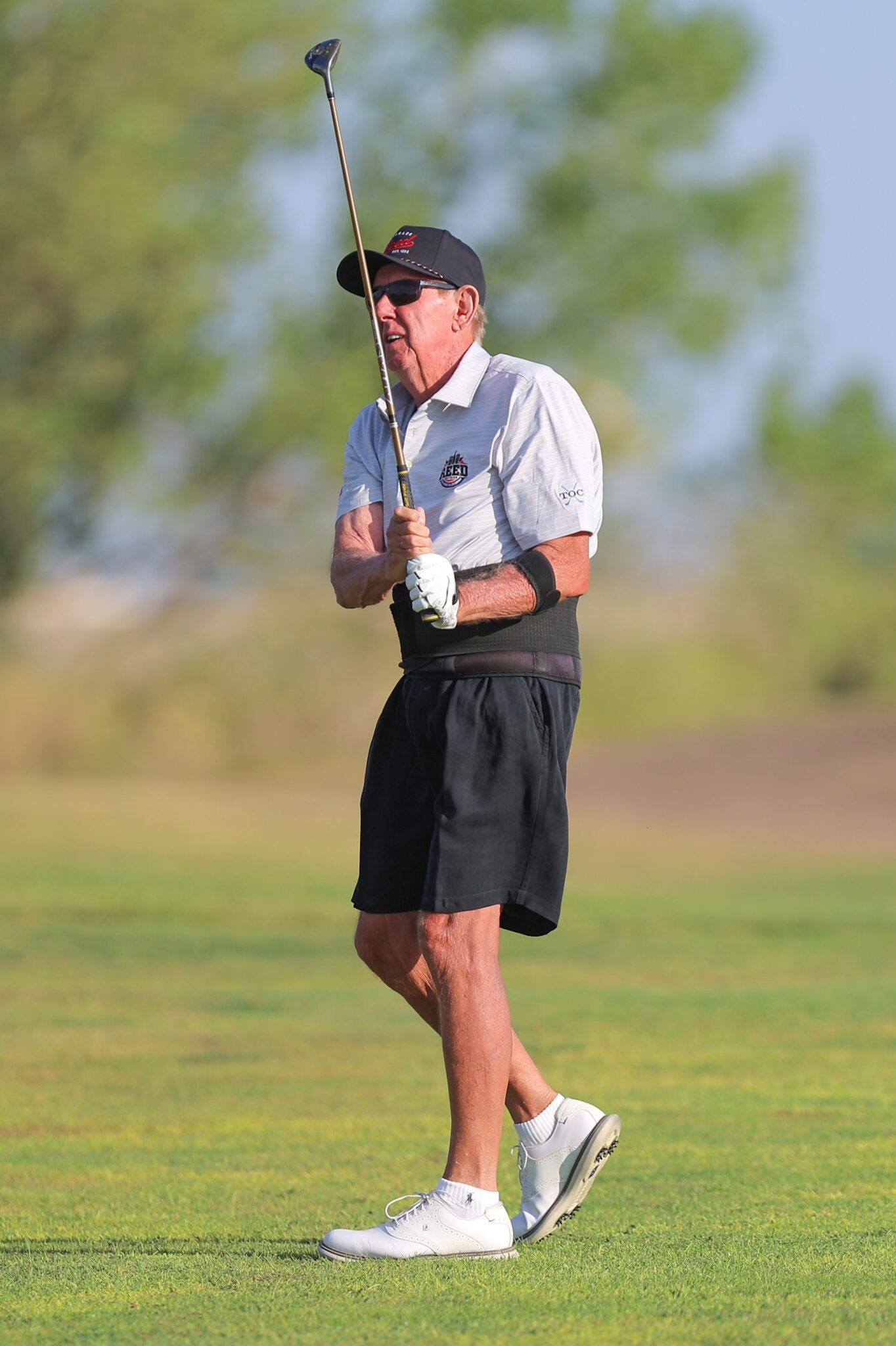20260815-CoorsTOC-Day3-004 | Press Pass Sports Older man in a white polo and black shorts, cap and sunglasses, preparing to swing a golf club on a sunny green golf course.