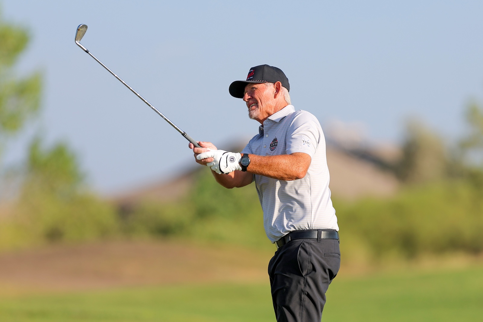 Older man in a cap and light polo shirt mid-swing with a golf club on a sunny golf course, wearing a glove and watch.