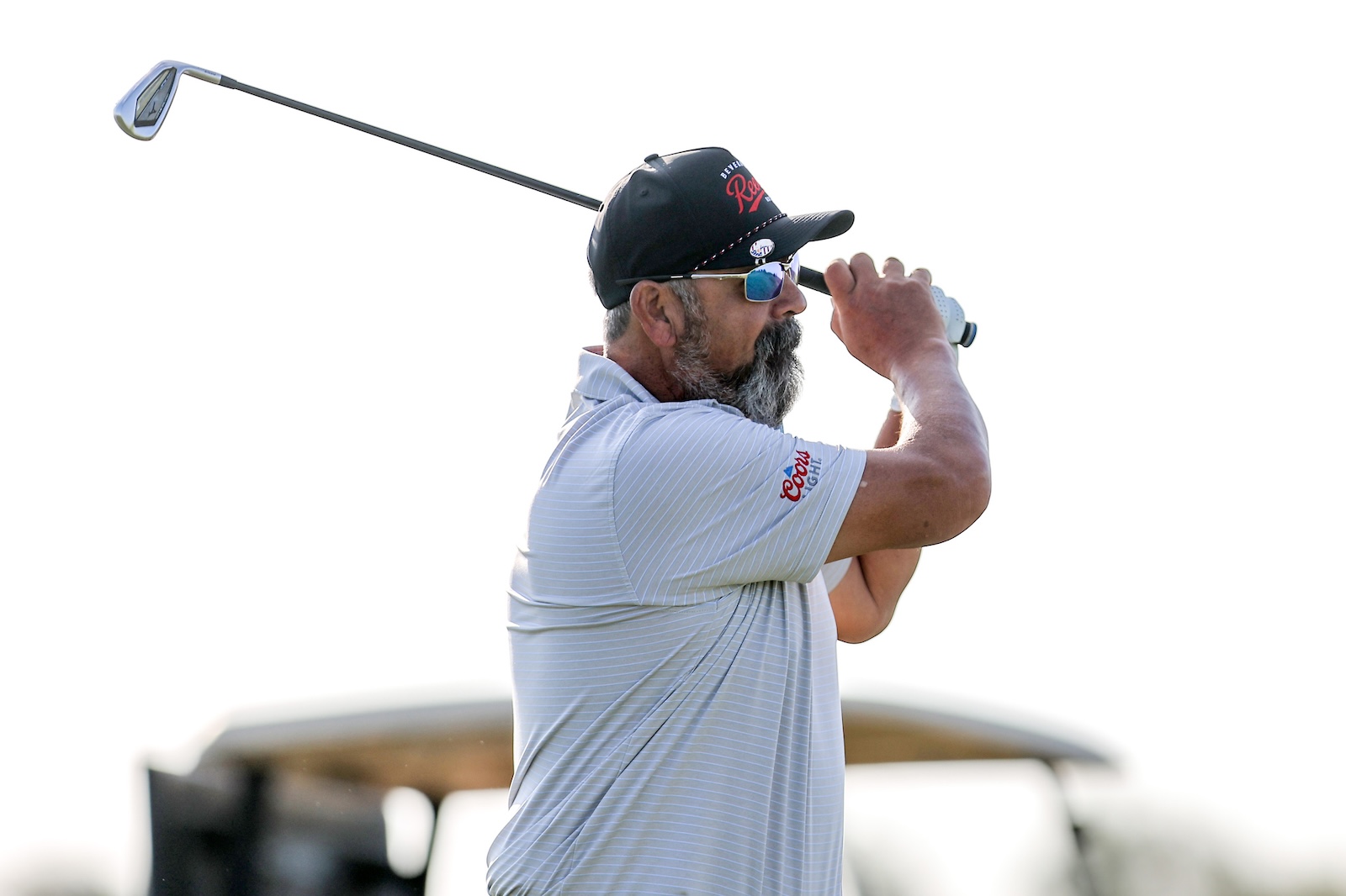 Bearded man in a dark cap and sunglasses mid-swing with a golf club, wearing a light striped polo with a Coors Light logo on the sleeve.