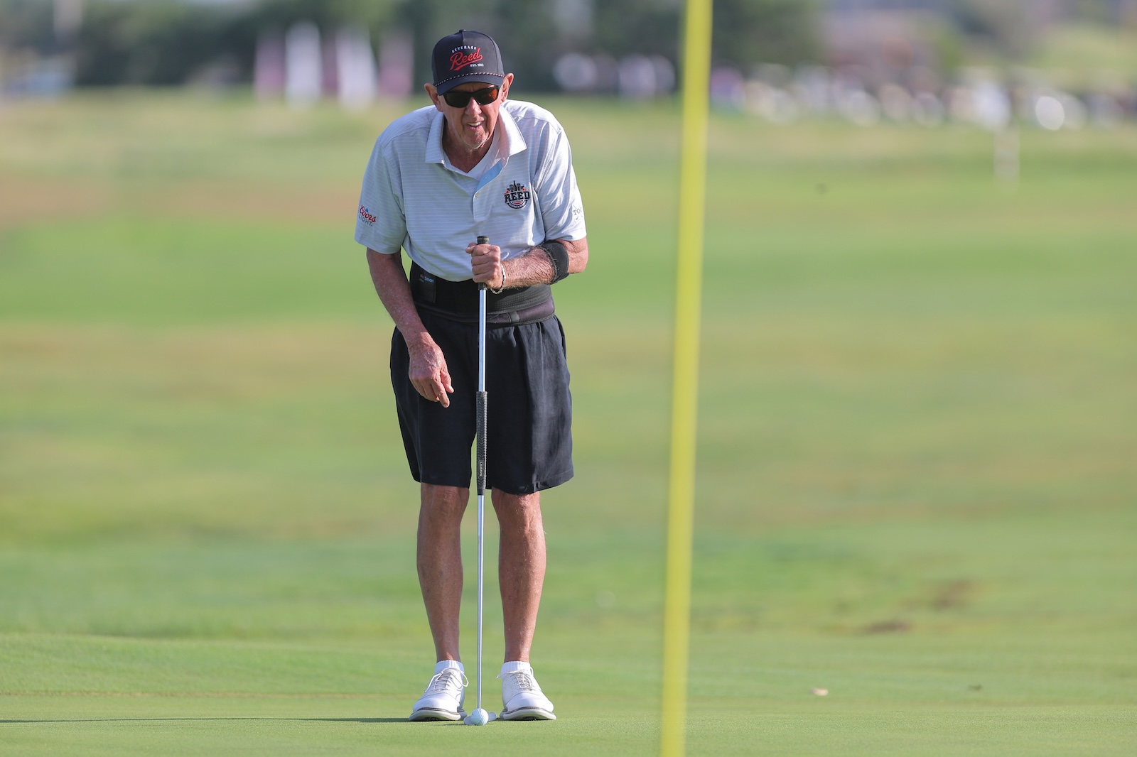An older man wearing a cap and sunglasses lines up a putt on a golf green, with a yellow flagstick in the background.