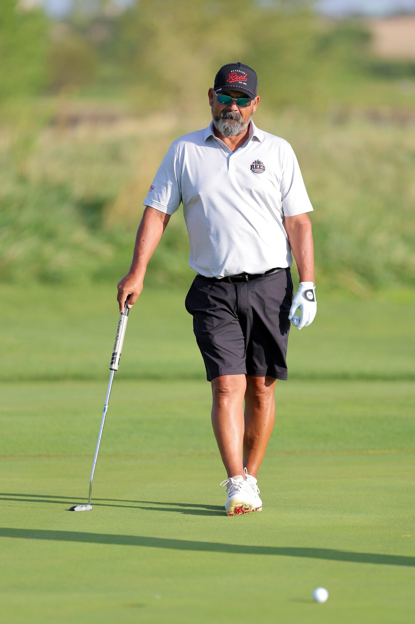 20260815-CoorsTOC-Day3-011 | Press Pass Sports Male golfer with gray beard in a white polo and dark cap standing on the putting green, preparing to putt with a putter and a ball nearby on the grass.