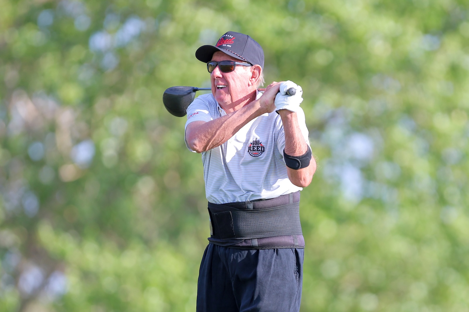 Older man in a polo shirt and cap mid-swing with a golf club on a sunny golf course, wearing sunglasses.