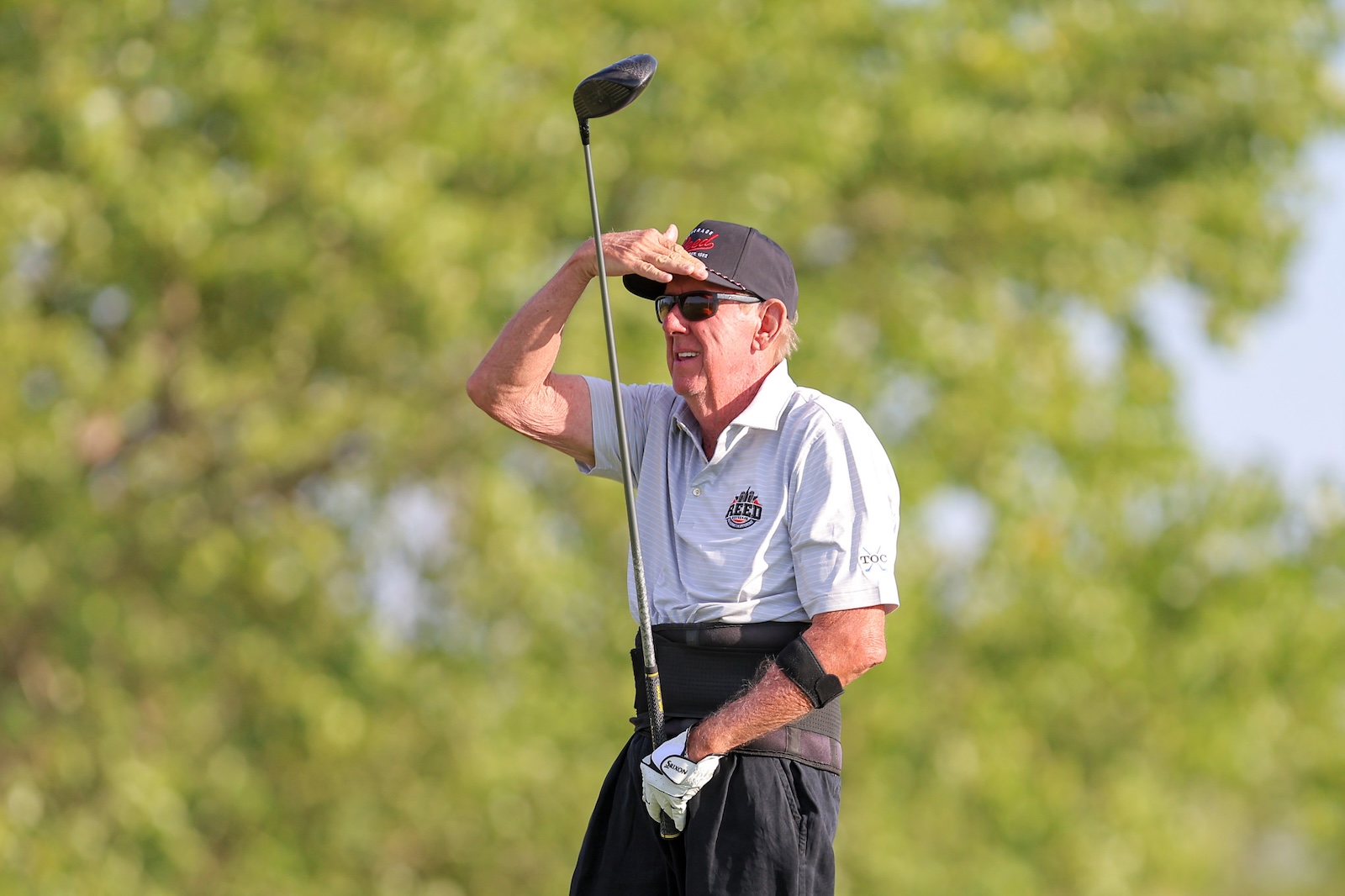 Older man in a cap and sunglasses shading his eyes with his hand while holding a golf club, on a sunny golf course area.