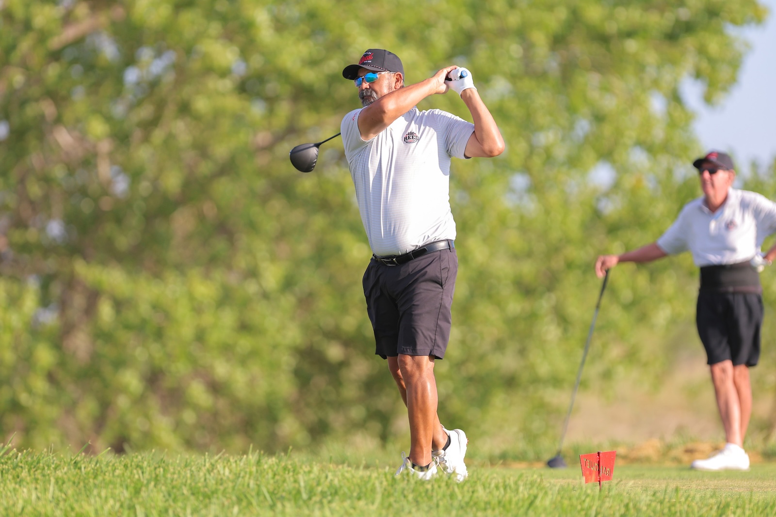 Adult man in a white polo and black shorts swinging a golf club on a sunny course, with another person observing in the background.