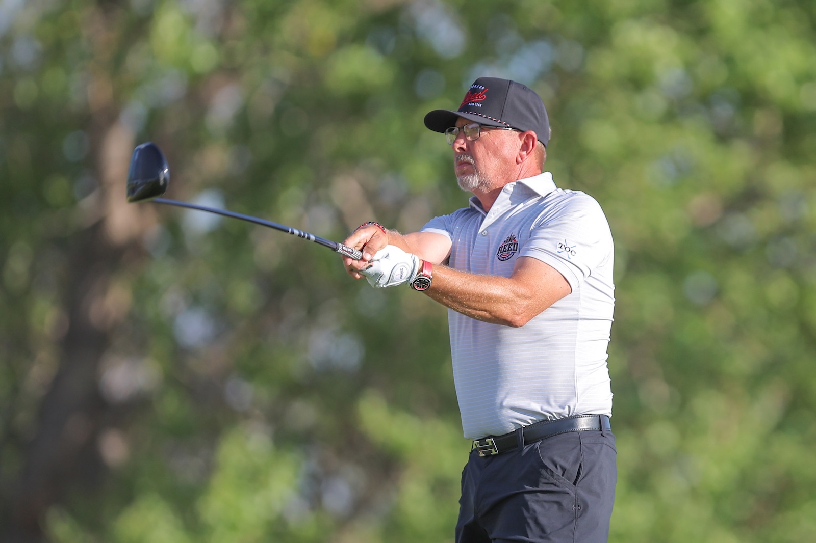 Man in a white polo and dark cap mid-swing with a driver on a sunny golf course.
