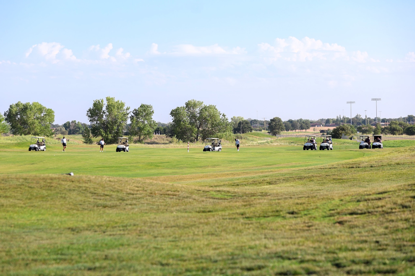 Wide view of a sunny golf course with players walking and golf carts along a green fairway.