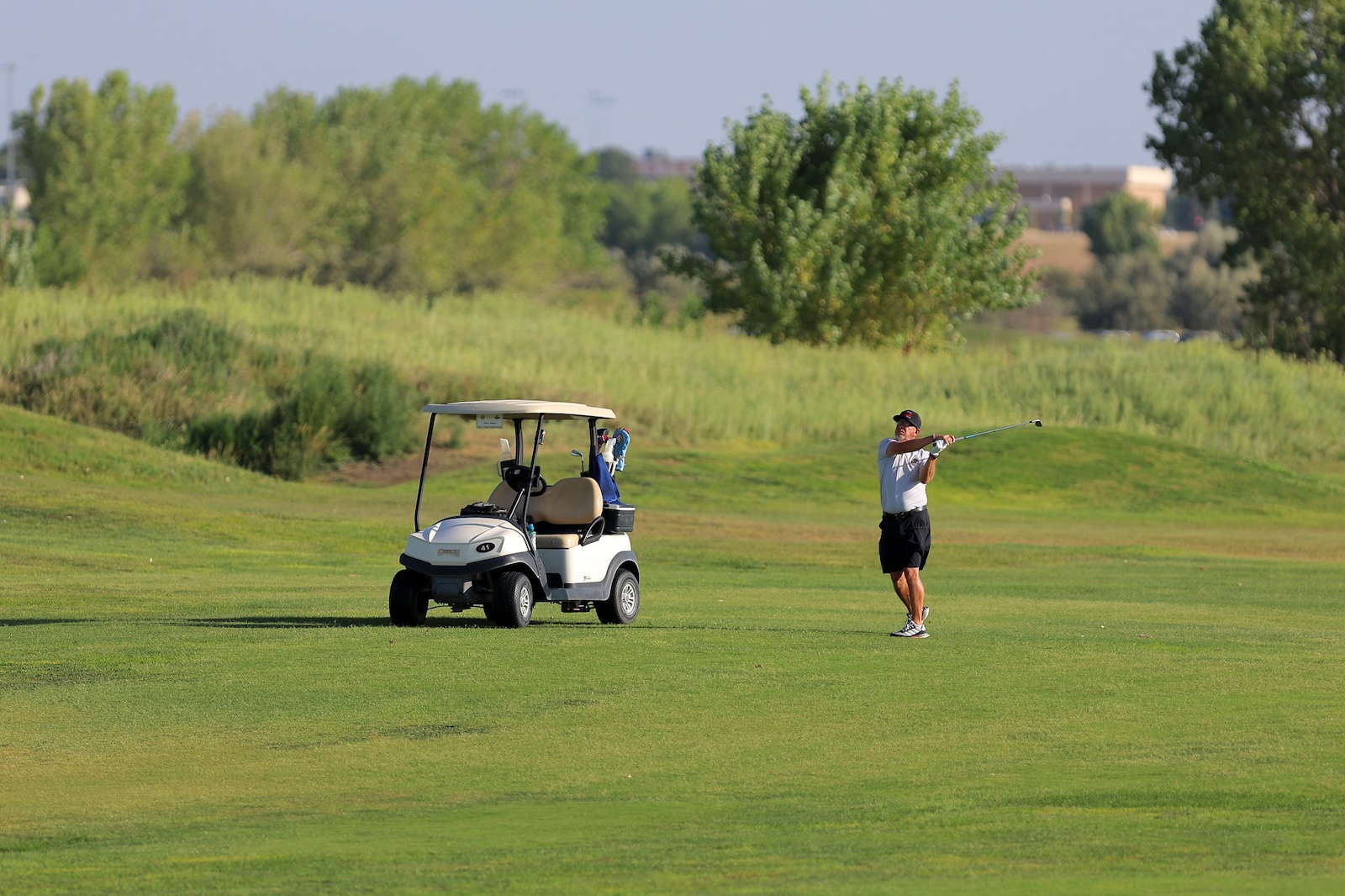 Golfer mid-swing on a sunny golf course beside a white golf cart.
