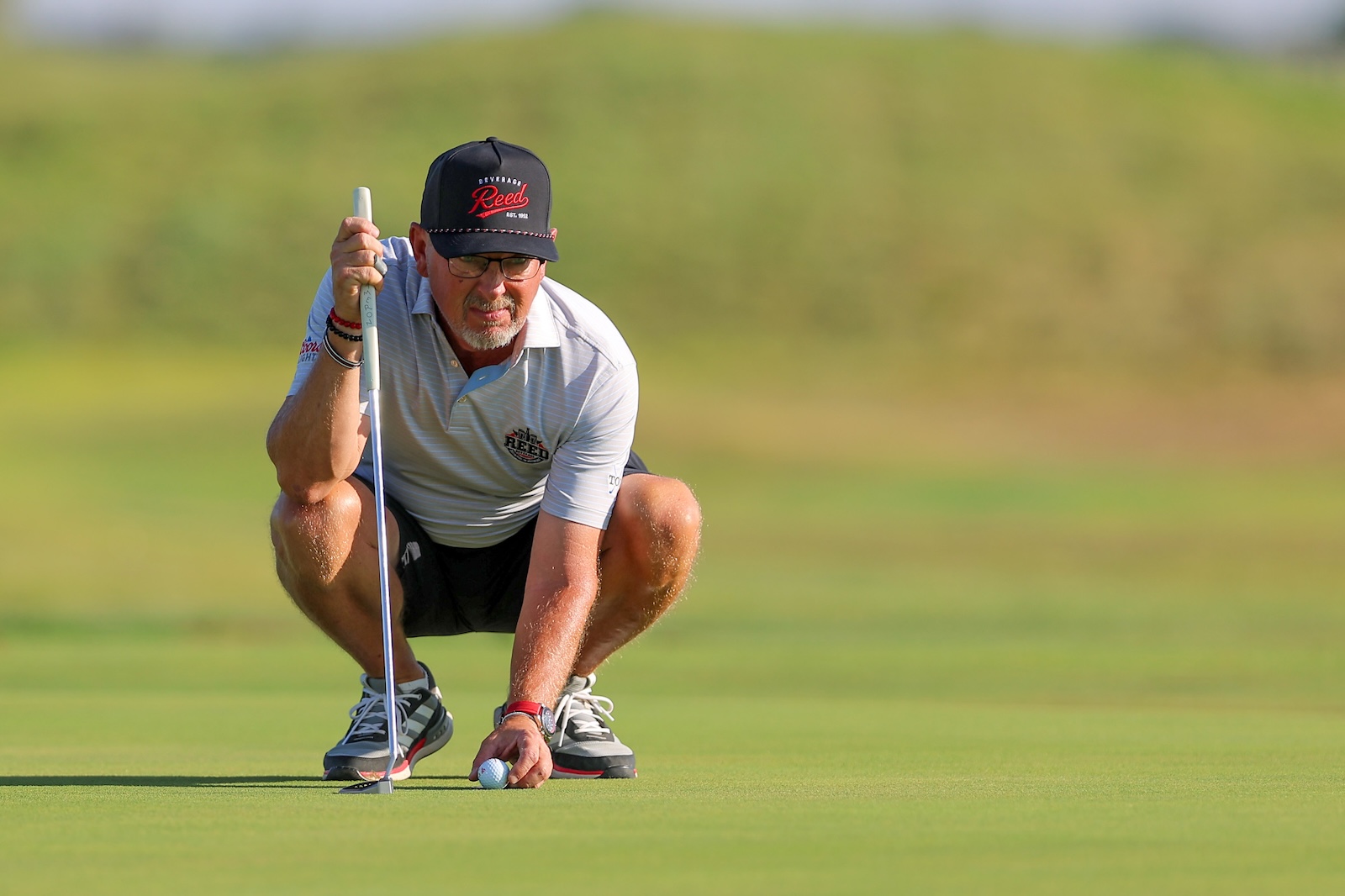 Older man in a gray polo crouches on a putting green lining up a shot with a golf flagstick nearby and a ball at his feet