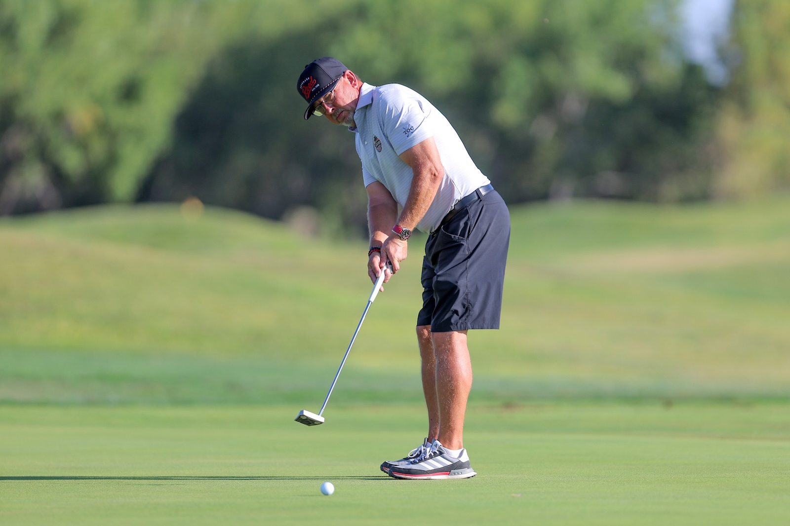 Male golfer in a white polo and dark shorts lines up a putt on a sunlit green golf course with trees in the background.