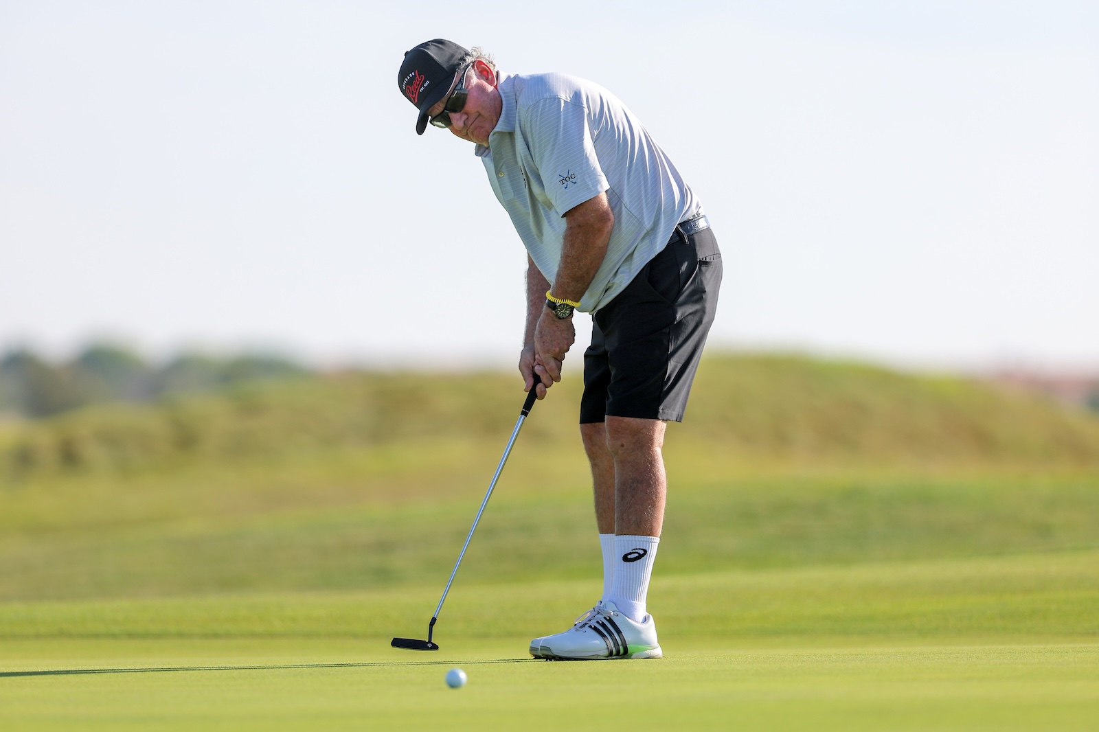 Man in a light polo and cap lining up a putt on a sunny golf green, wearing sunglasses and a watch.