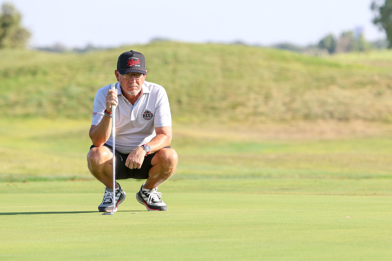 Older man crouching on a golf green, holding a putter and preparing to line up a putt, wearing a white polo and cap.