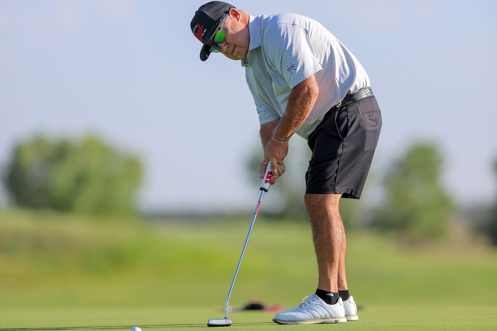 A man wearing a cap and sunglasses lines up a putt on a golf green, holding a putter with both hands.