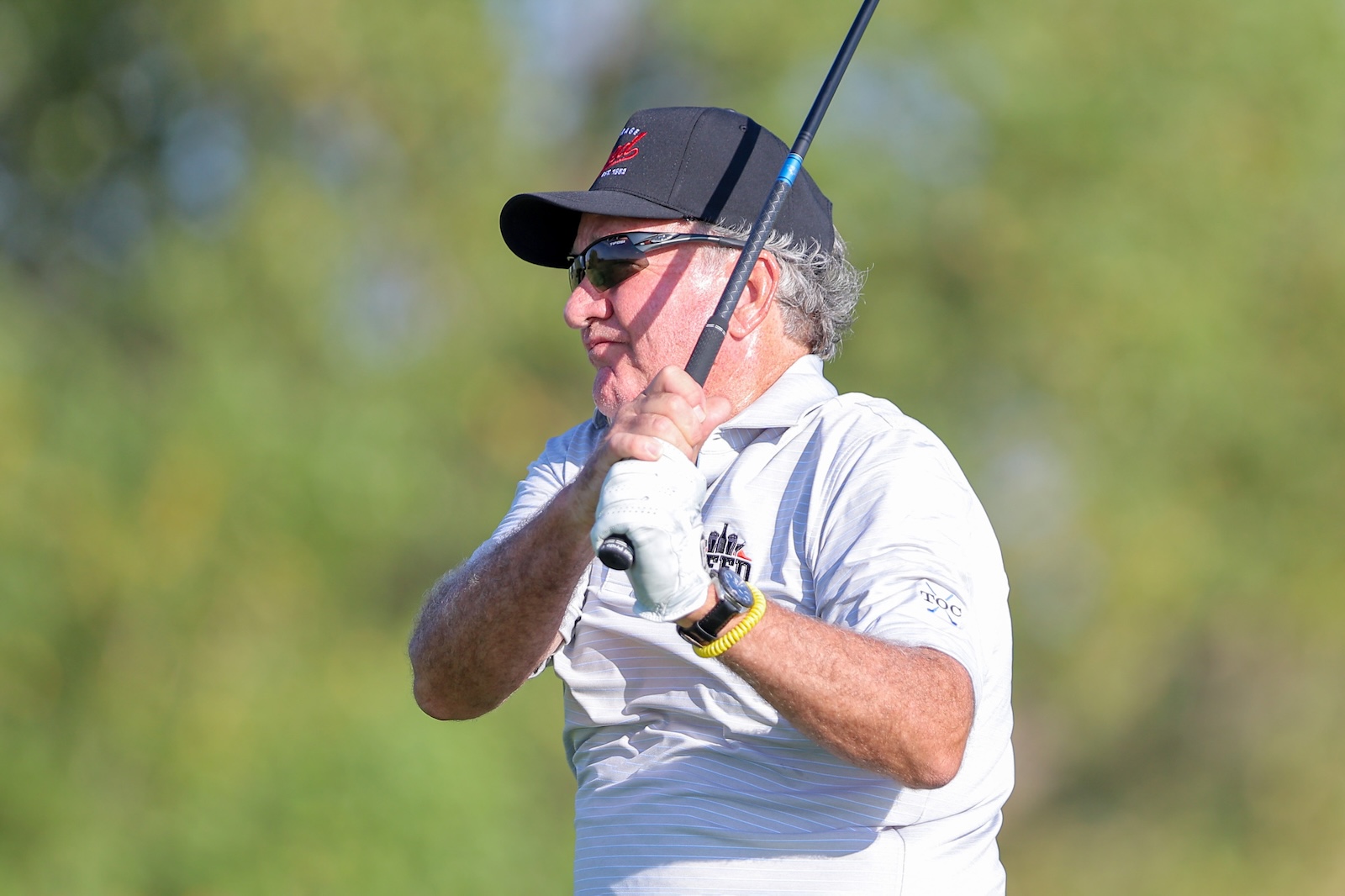 Male golfer wearing a black cap and sunglasses mid-swing with a golf club outdoors.