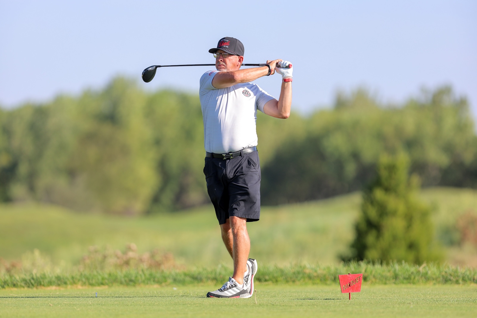 Older man in a gray polo and cap swinging a golf club on a sunny course, with a red tee marker nearby.