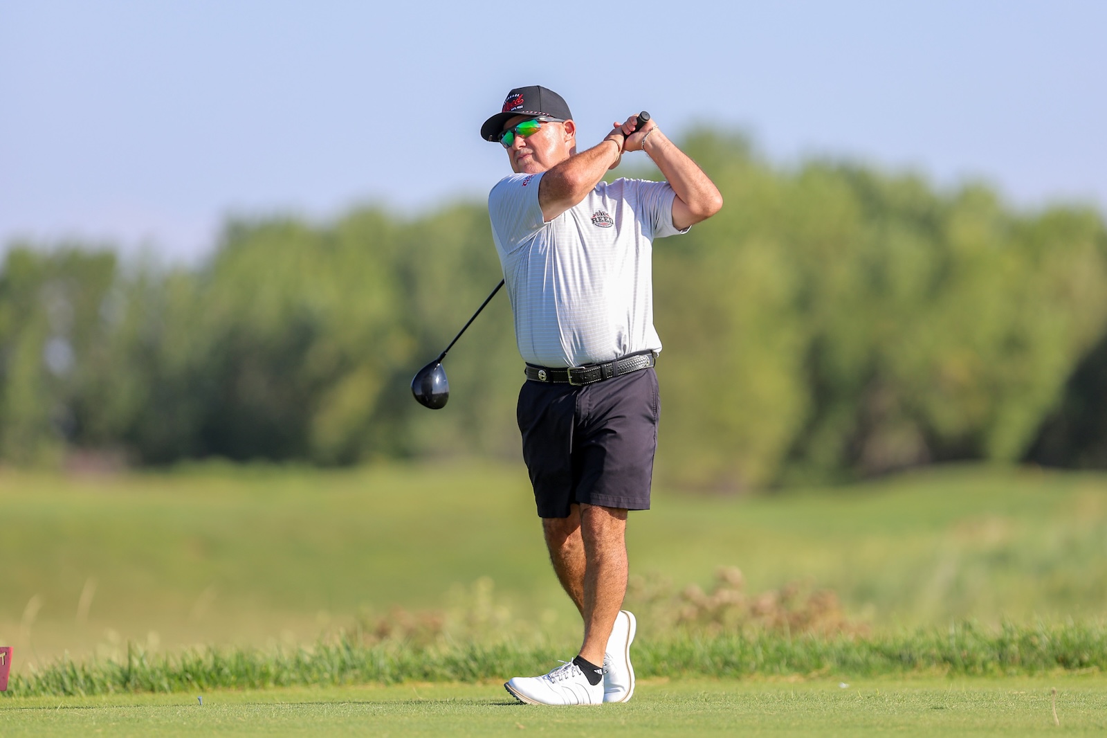 Man in a light gray polo and dark shorts mid-swing on a golf course, wearing a cap and sunglasses as he hits the ball.