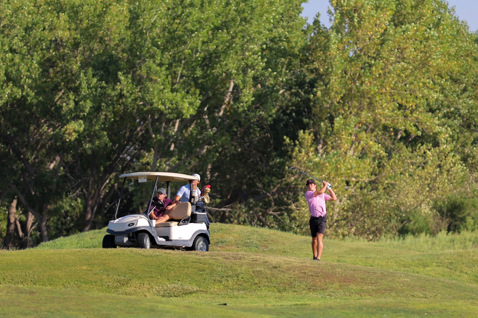 Golfer in a pink polo swinging a golf club on a sunny course, with a white golf cart and three passengers nearby under green trees.
