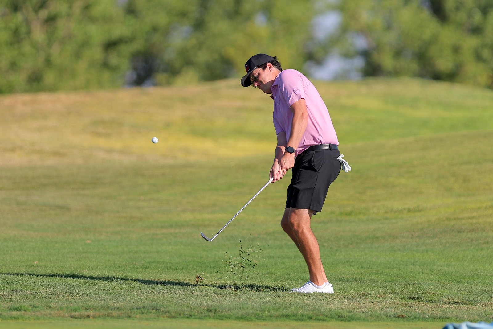 A man in a pink polo and black cap drives a golf ball on a green fairway, mid-swing with a clubsmen stance.