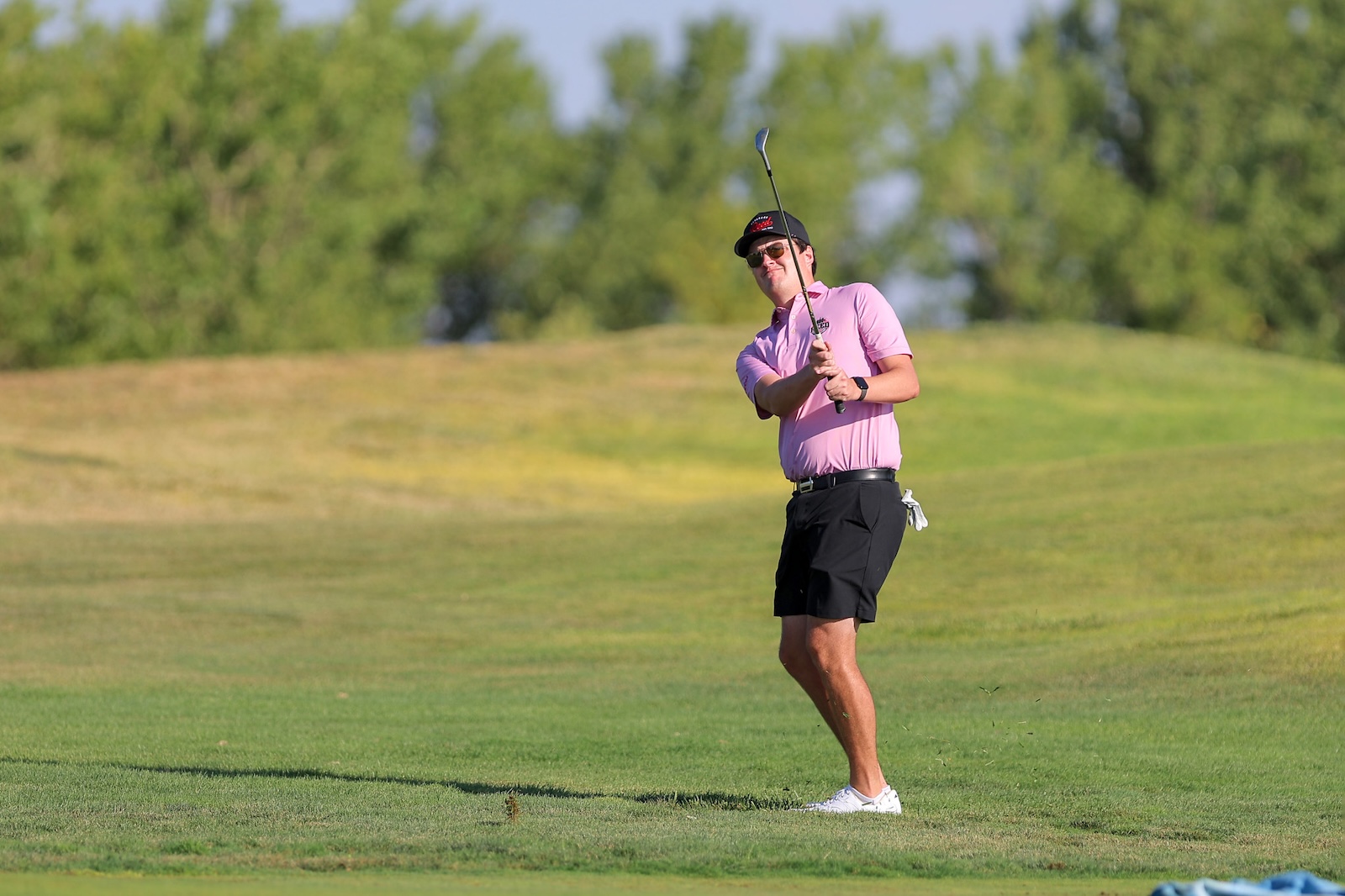 Male golfer in a pink polo and black shorts mid-swing on a sunny golf course, holding a golf club high above his shoulder.