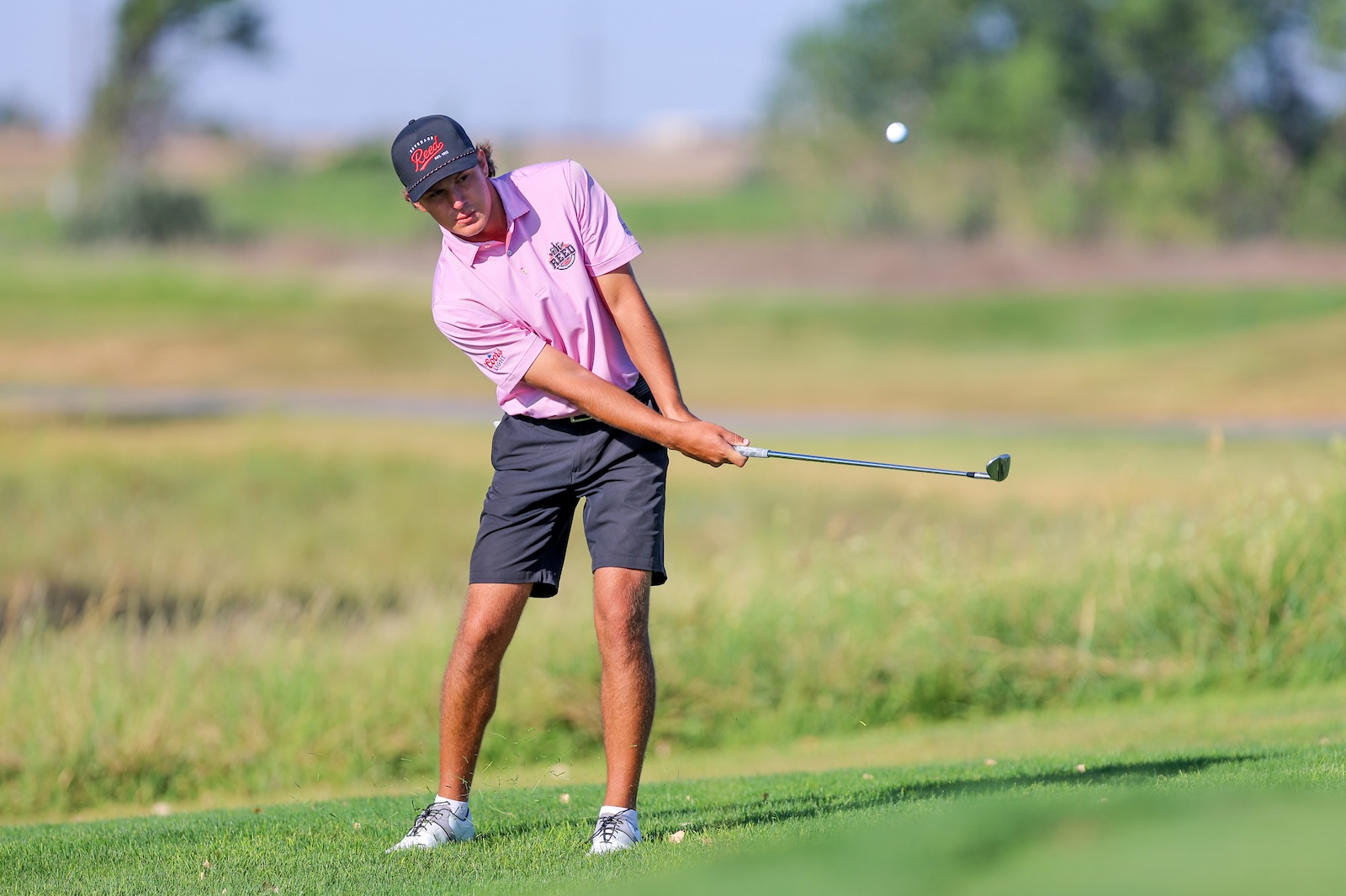 Male golfer in a pink polo and black cap swinging a club on a sunny golf course.