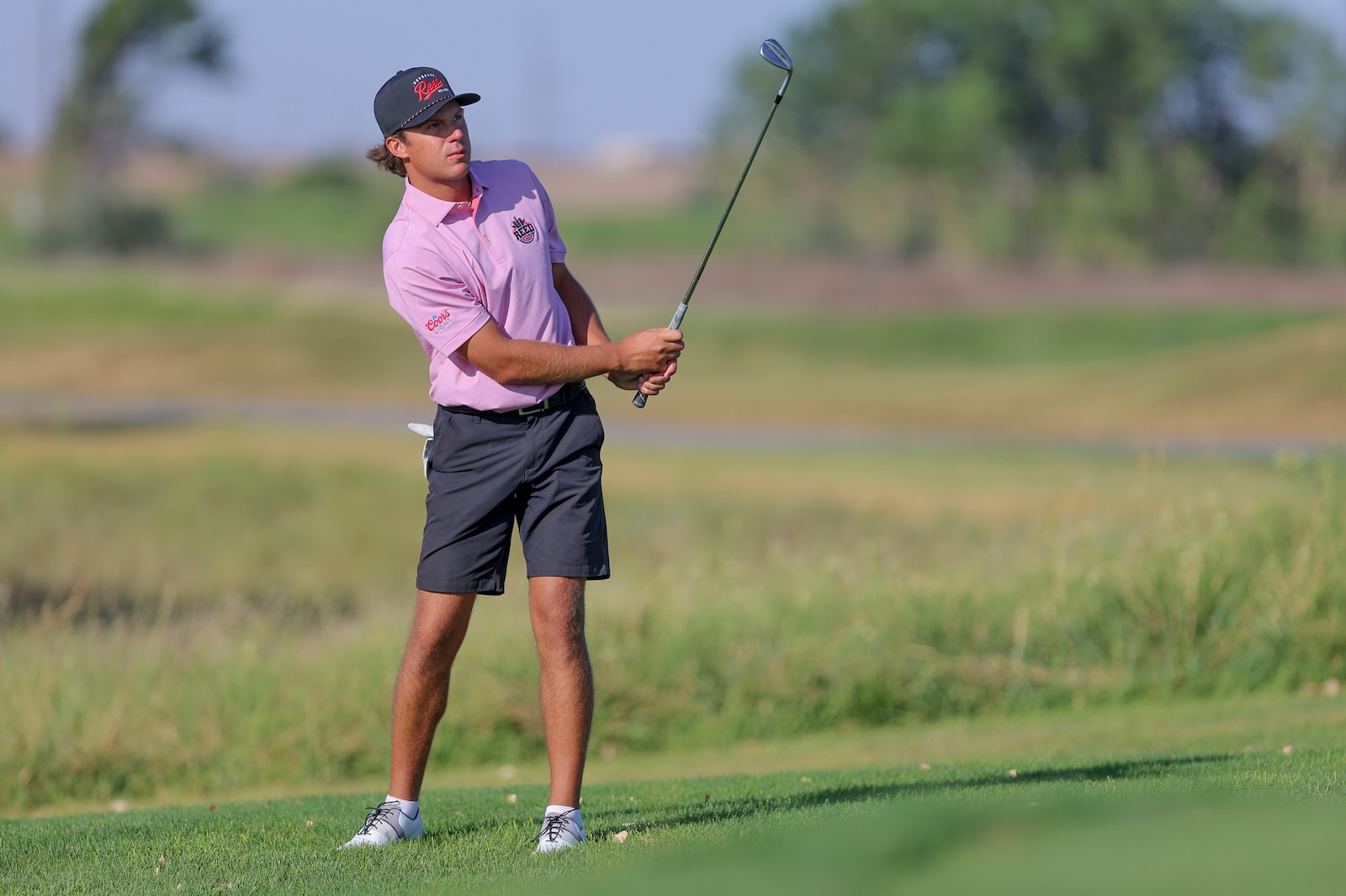 Male golfer in a pink polo shirt and dark shorts mid-swing on a sunny golf course.