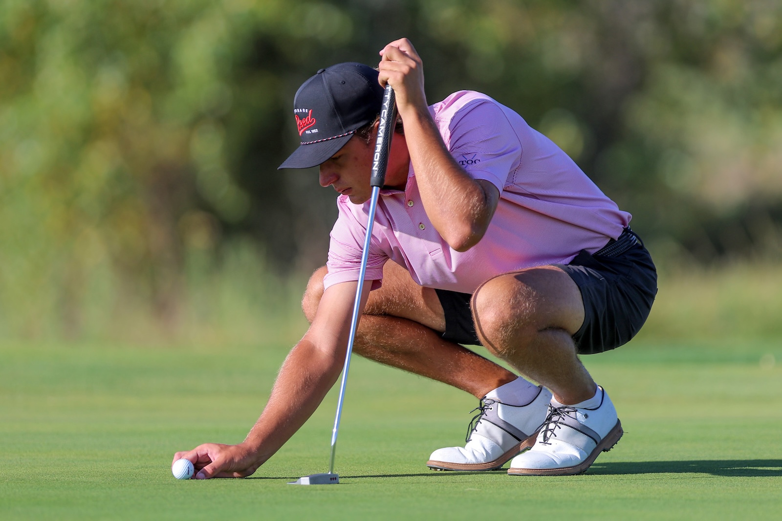 Golfer in a pink polo crouches on the green, lining up a putt with a club.