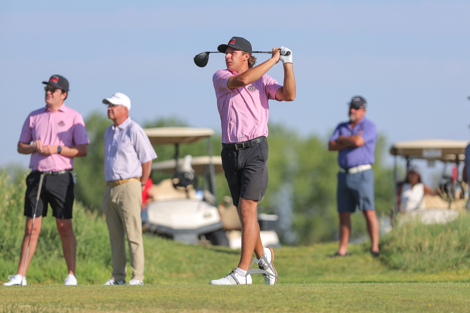 Golfer in pink shirt mid-swing on a tee while spectators watch on a sunny course.