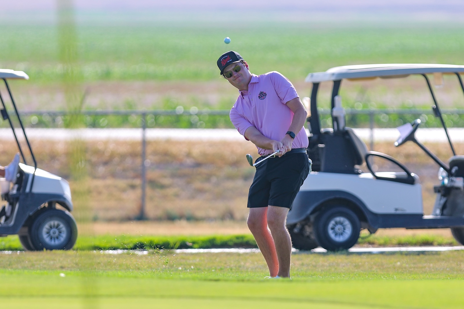 Man in a pink polo and cap mid-swing on a sunny golf course, ball in the air with golf carts in the background