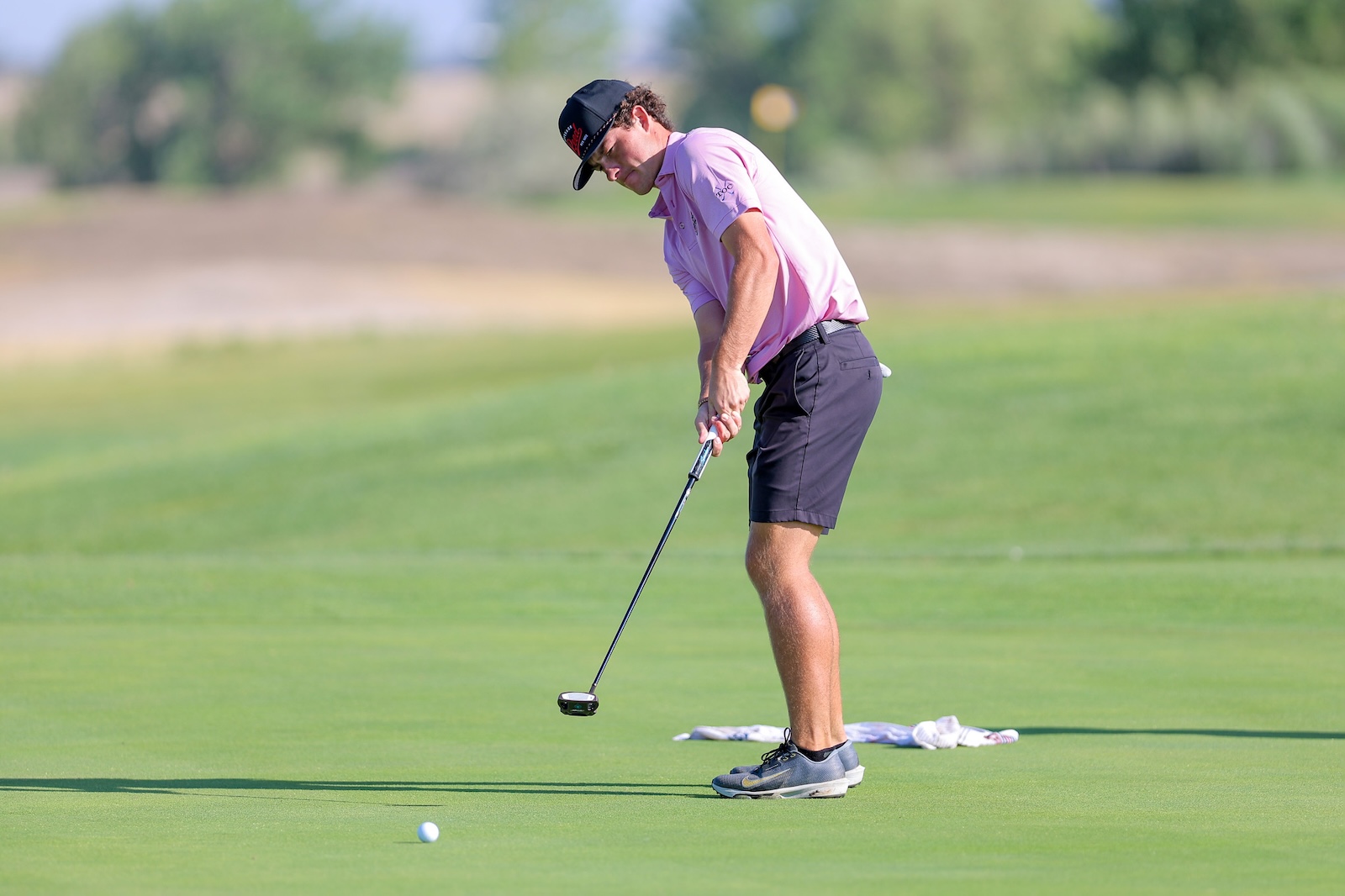 Golfer in a pink polo and black cap lining up a putt on a lush green putting surface, with a white golf ball nearby.
