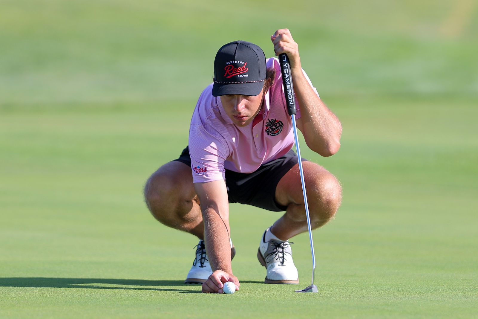 Golfer in a pink polo crouches to line up a putt on the green, ball near the hole with a putter in hand.