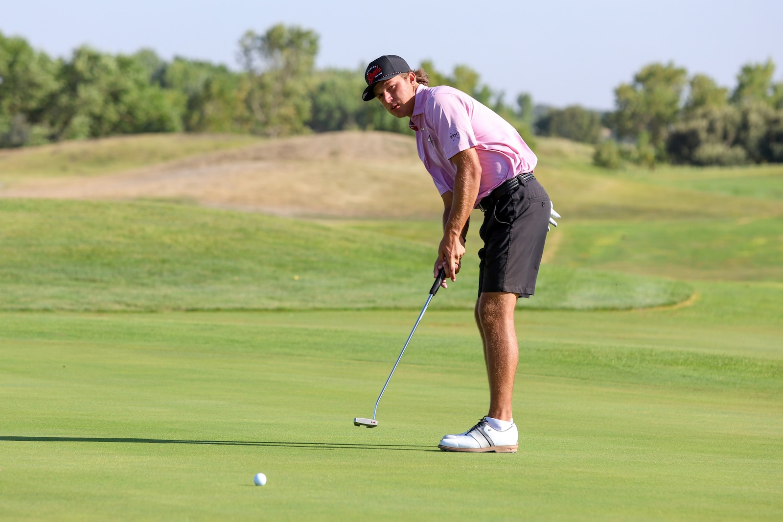 Golfer in a pink polo shirt and black cap preparing a putt on a sunny green golf course.