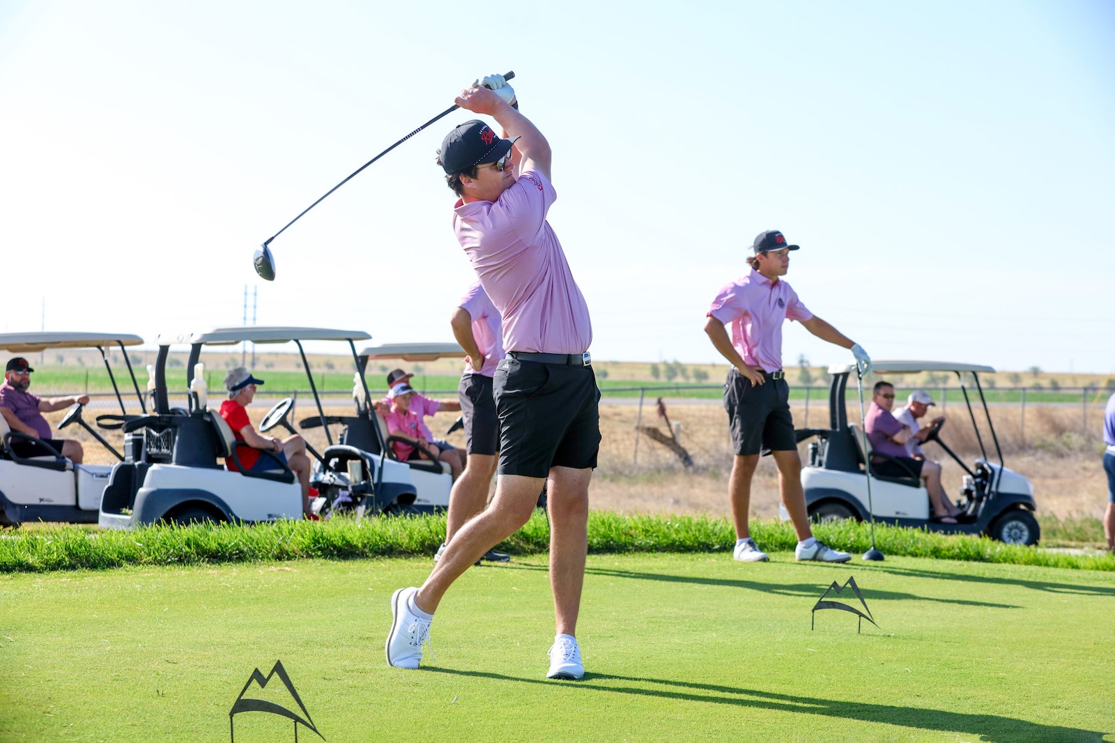 A man in a pink polo swings a golf club on a sunny course while others watch from a line of golf carts in the background.