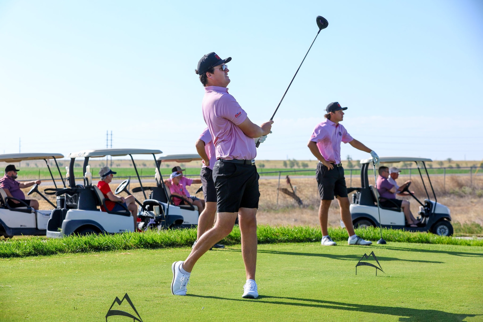 Man in pink polo swinging a golf club on a sunny course, with teammates and golf carts in the background.