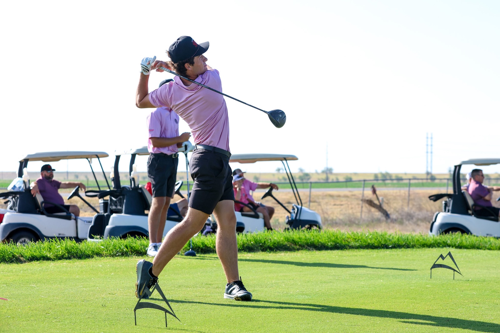 Golfer in pink shirt and cap swings a driver on a sunny golf course, carts and players in the background