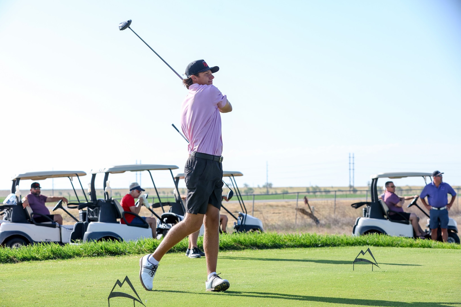 Golfer in pink shirt mid-swing on a sunny golf course with carts and spectators in the background.