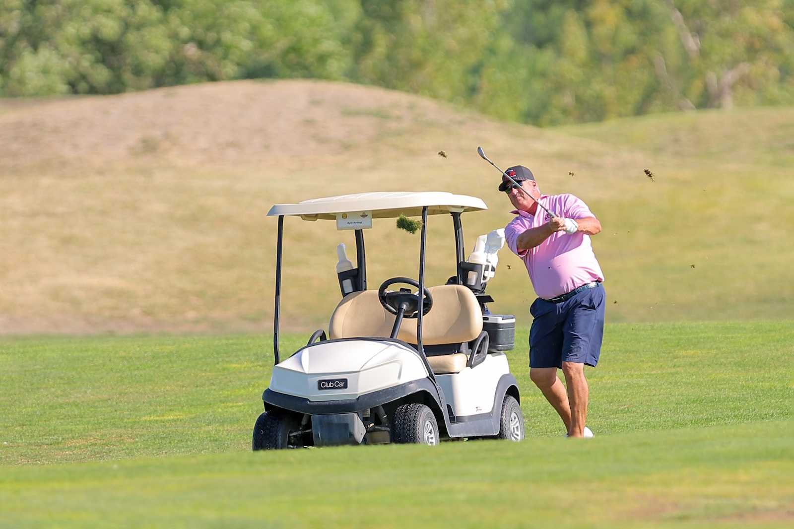 A man in a pink polo shirt swings a golf club next to a beige golf cart on a sunny golf course.