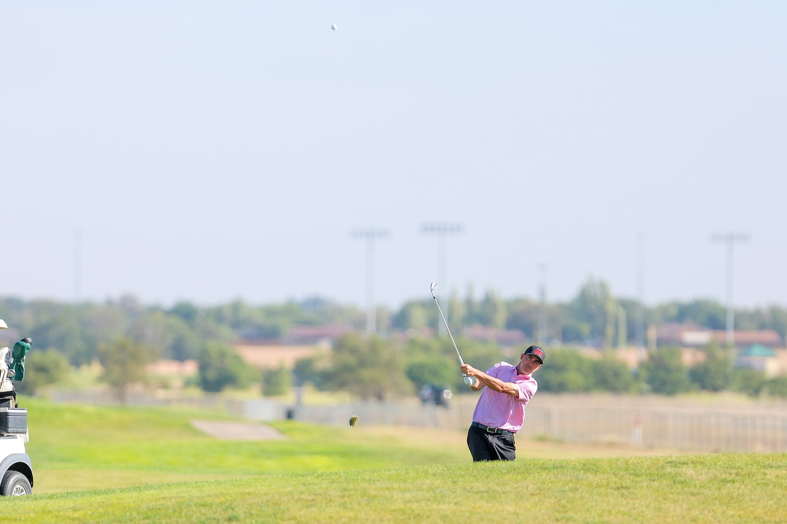 Golfer in a pink shirt takes a powerful swing on a sunny golf course, ball in midair with a cart on the left.