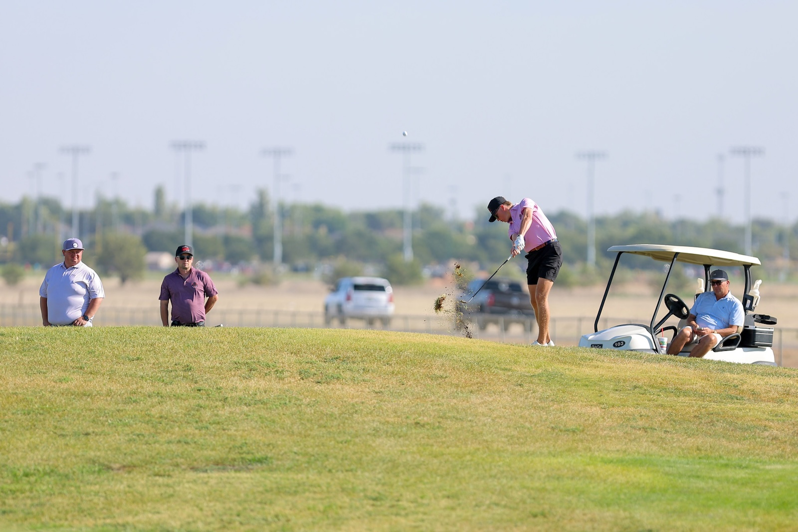 Golfer in pink shirt and black shorts hits from a sand bunker while a ball flies, with spectators and a golf cart nearby on a sunny course.