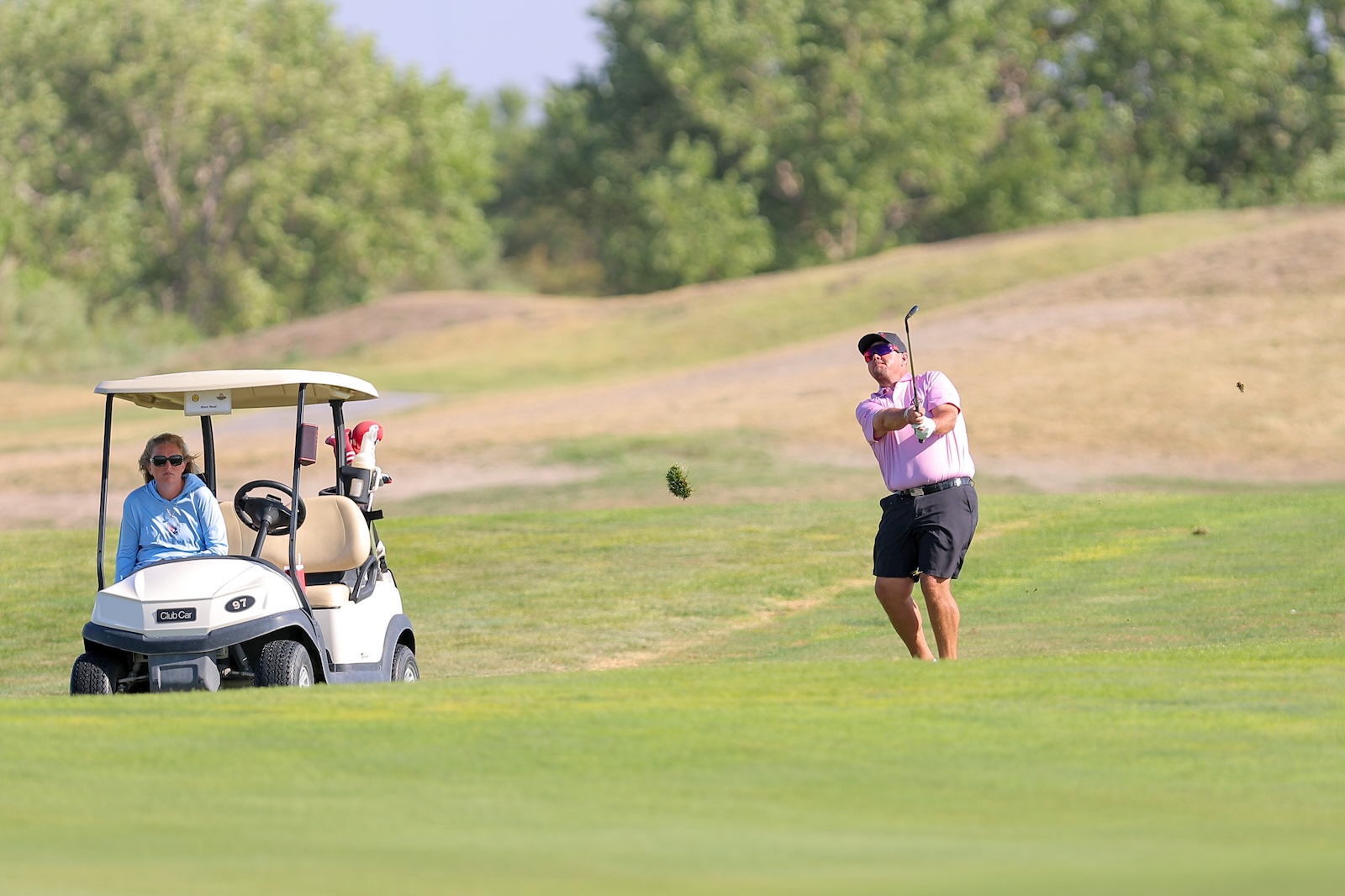Man in a pink shirt swings a golf club on a sunny course; a woman sits in a white golf cart nearby.
