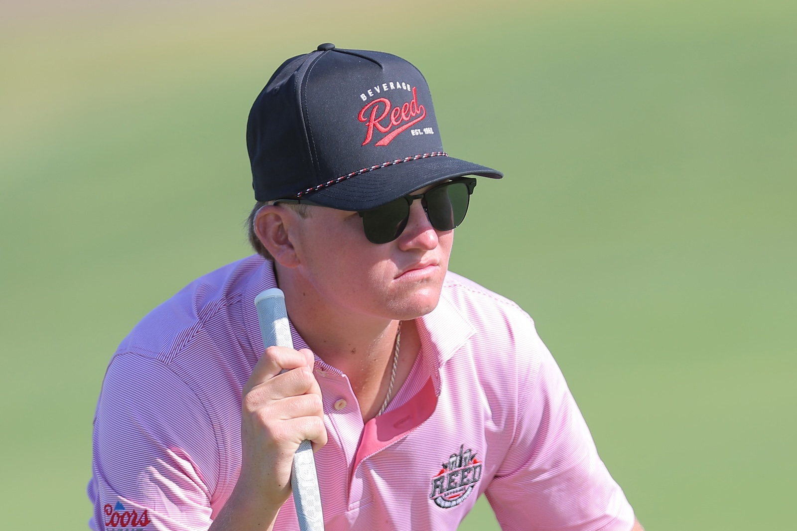 Man in a pink polo and black cap with sunglasses holding a golf club on a sunny course.