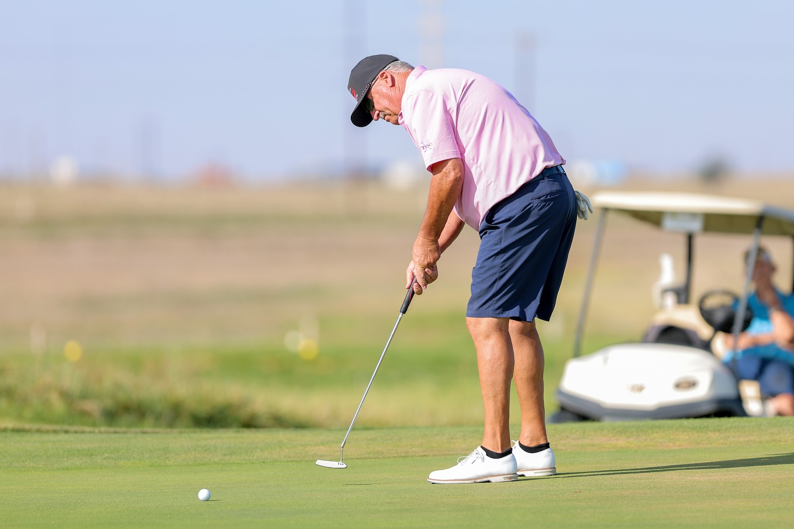 Older man in a pink polo and navy shorts puts on a sunny golf green, with a cart in the background.