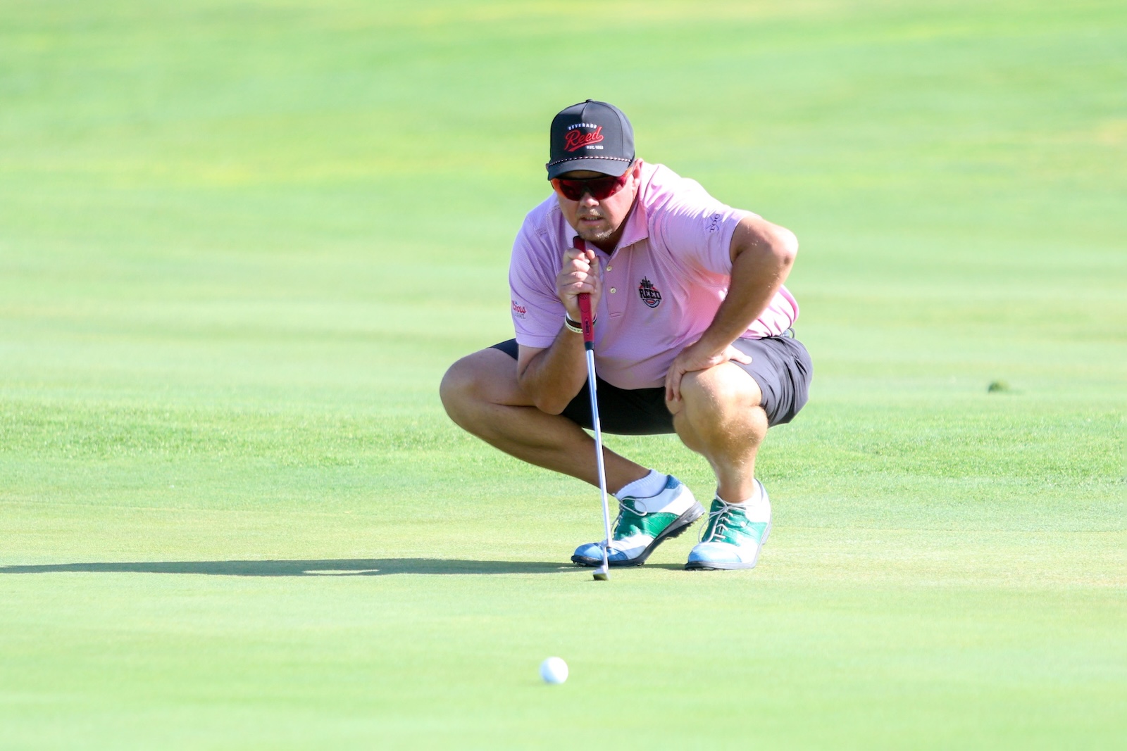 Golfer crouching on the green, lining up a putt with a pink polo, cap, and teal shoes.