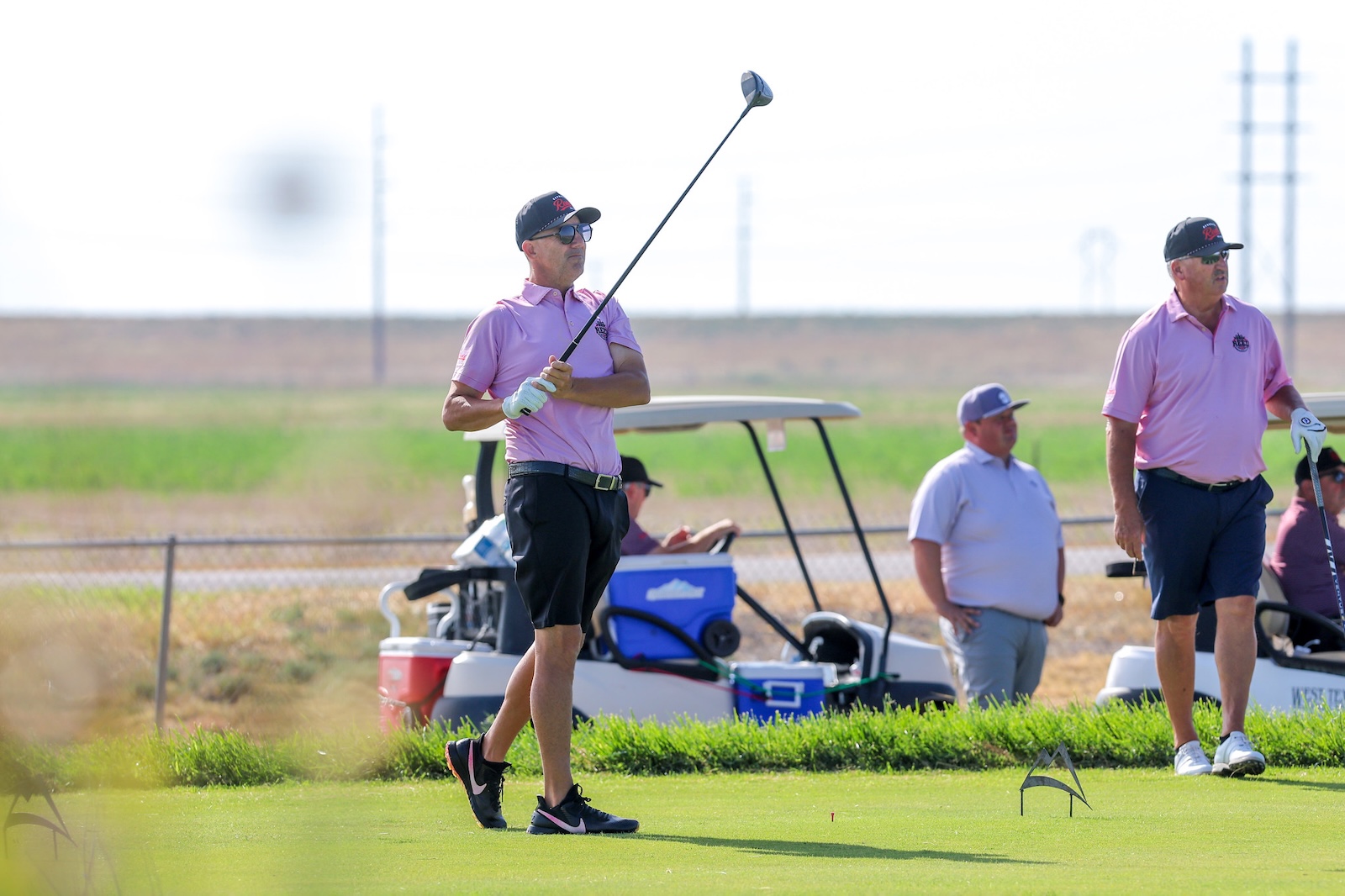 Man in a pink polo shirt swings a golf club on a sunny course, with teammates and a golf cart in the background.