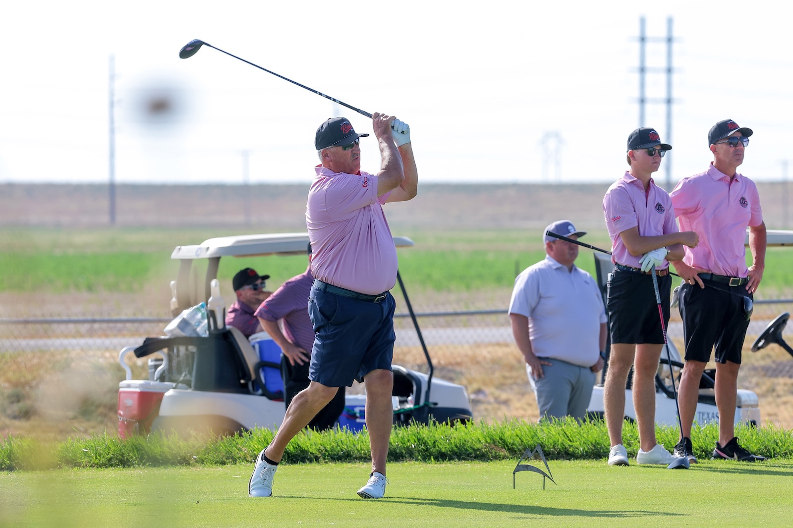 A man in a pink polo swings a golf club on a sunny course while fellow players watch nearby.