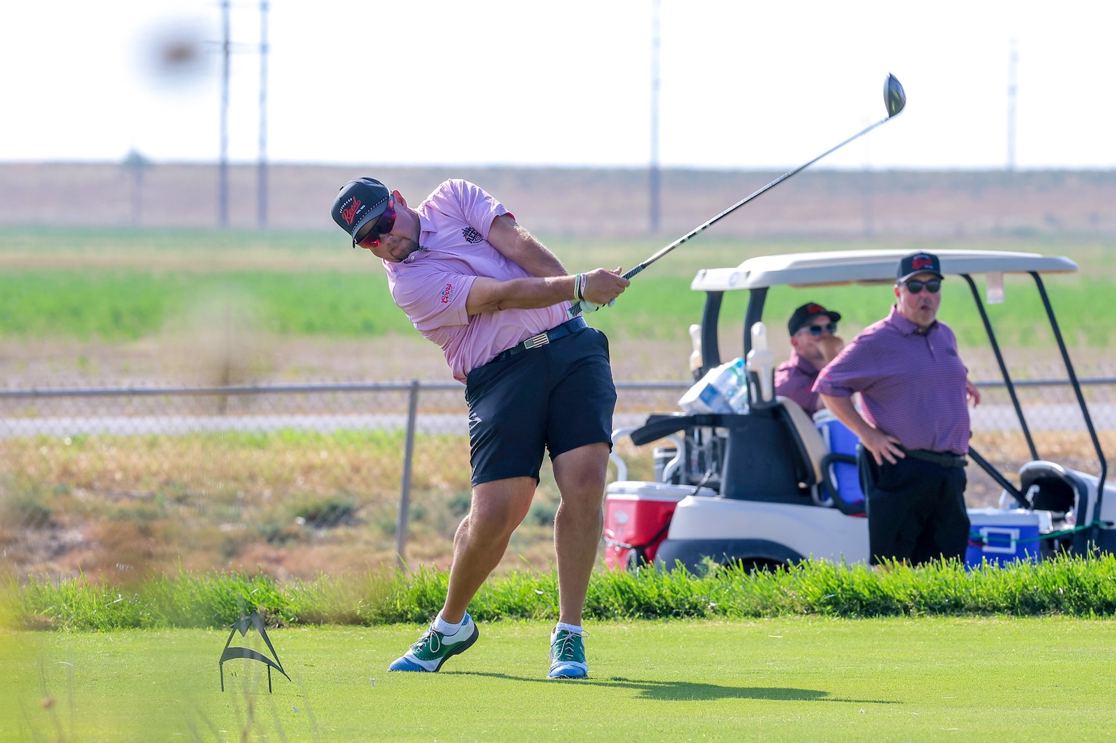 Golfer in a pink polo mid-swing on a sunny golf course with a cart and two spectators in the background