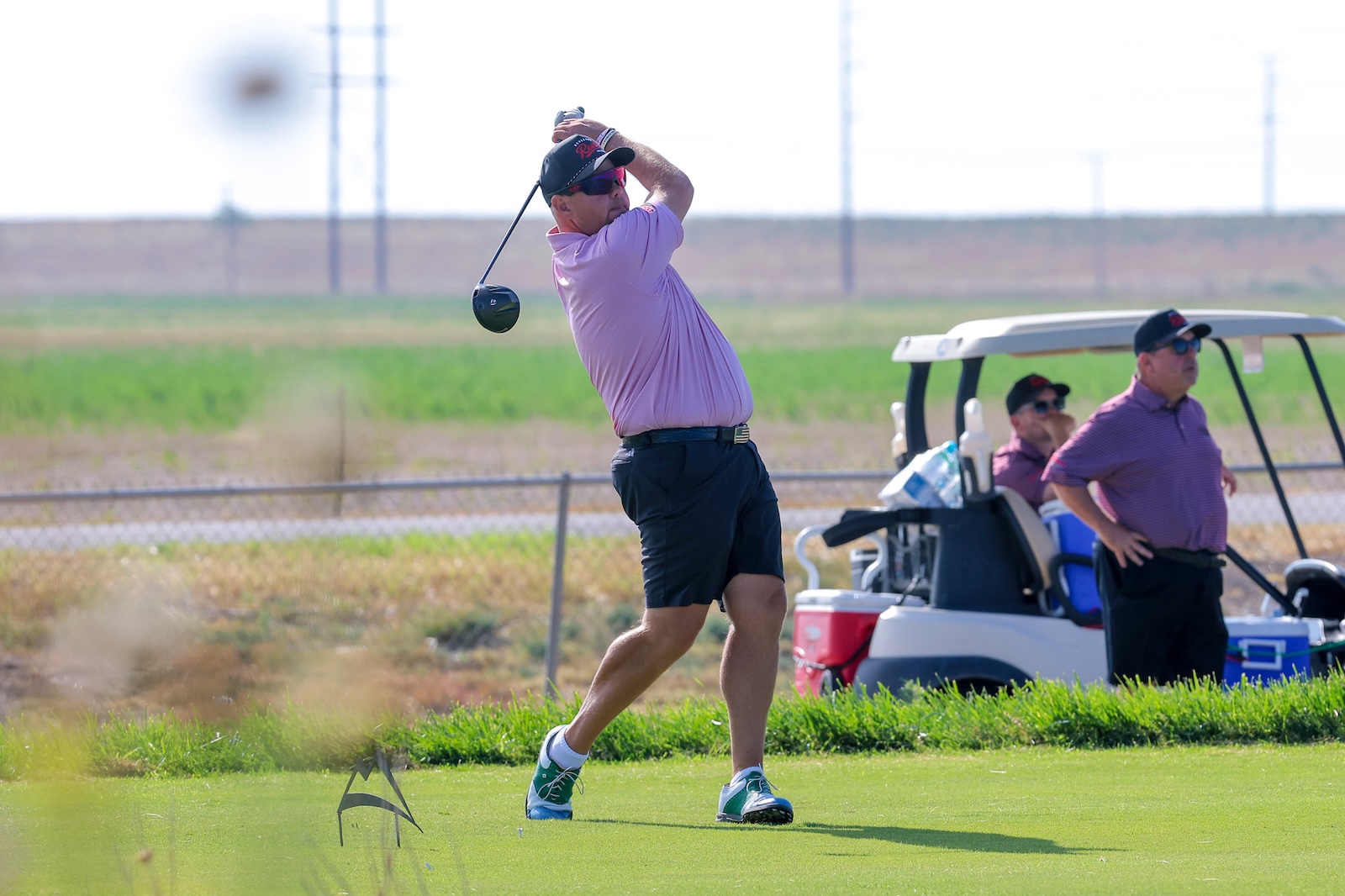 Golfer in a pink shirt and cap mid-swing on a sunny golf course, with two people by a golf cart in the background.