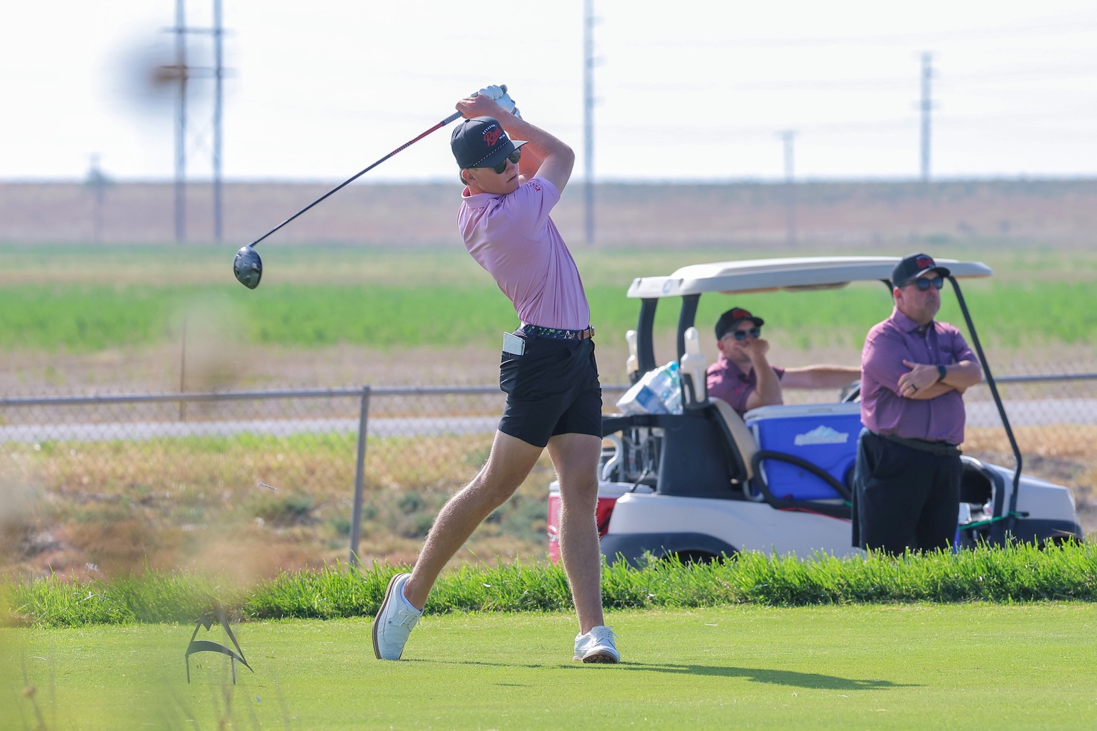 Golfer in a pink polo swings a driver on a grassy course with a cart and two spectators in the background.