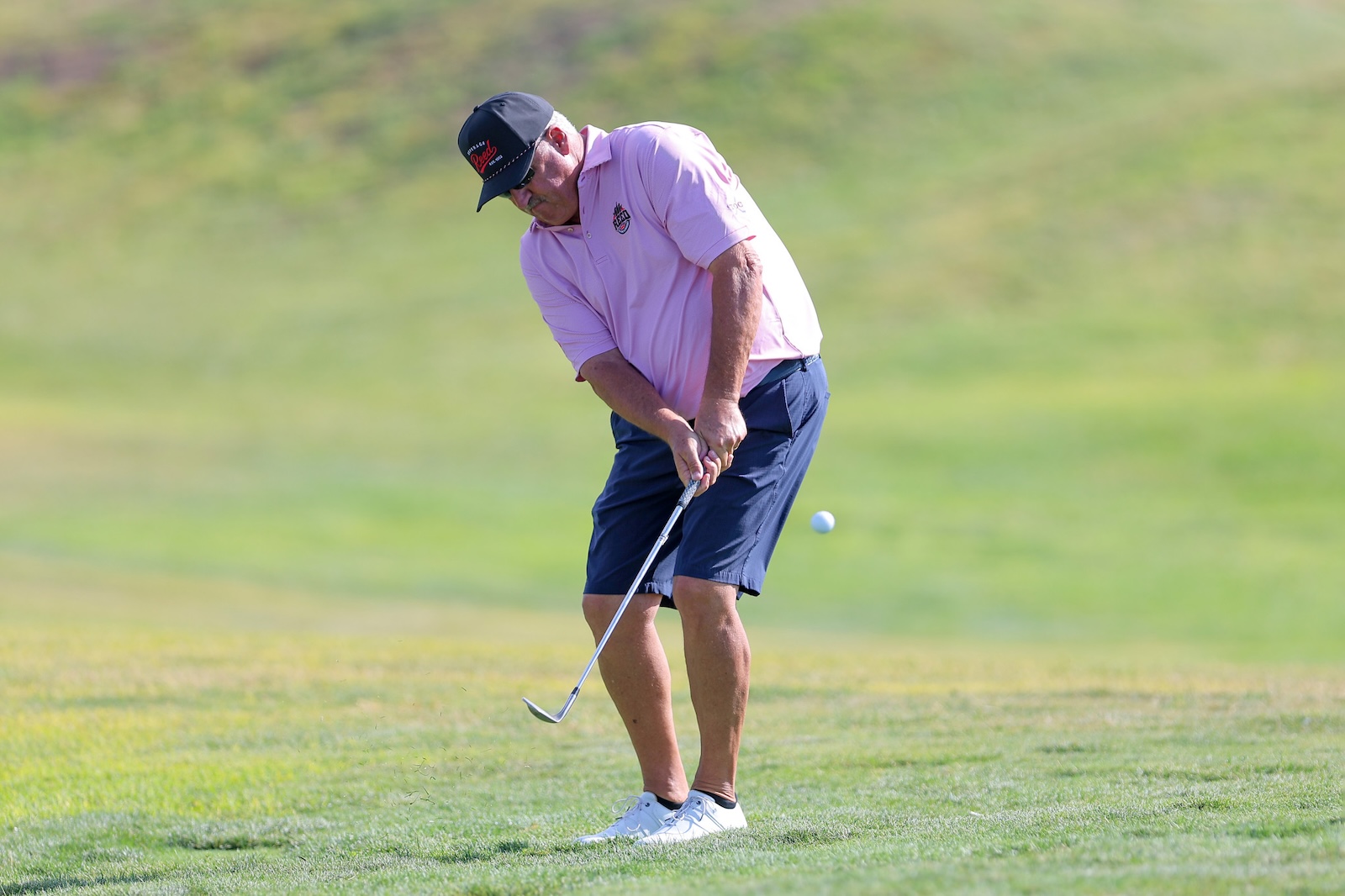 Older man in a pink polo and navy shorts taking a golf swing on a sunny green course.