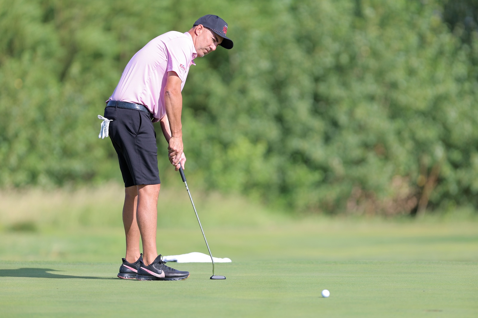Golfer in a pink polo and black shorts lines up a putt on the sunny green.
